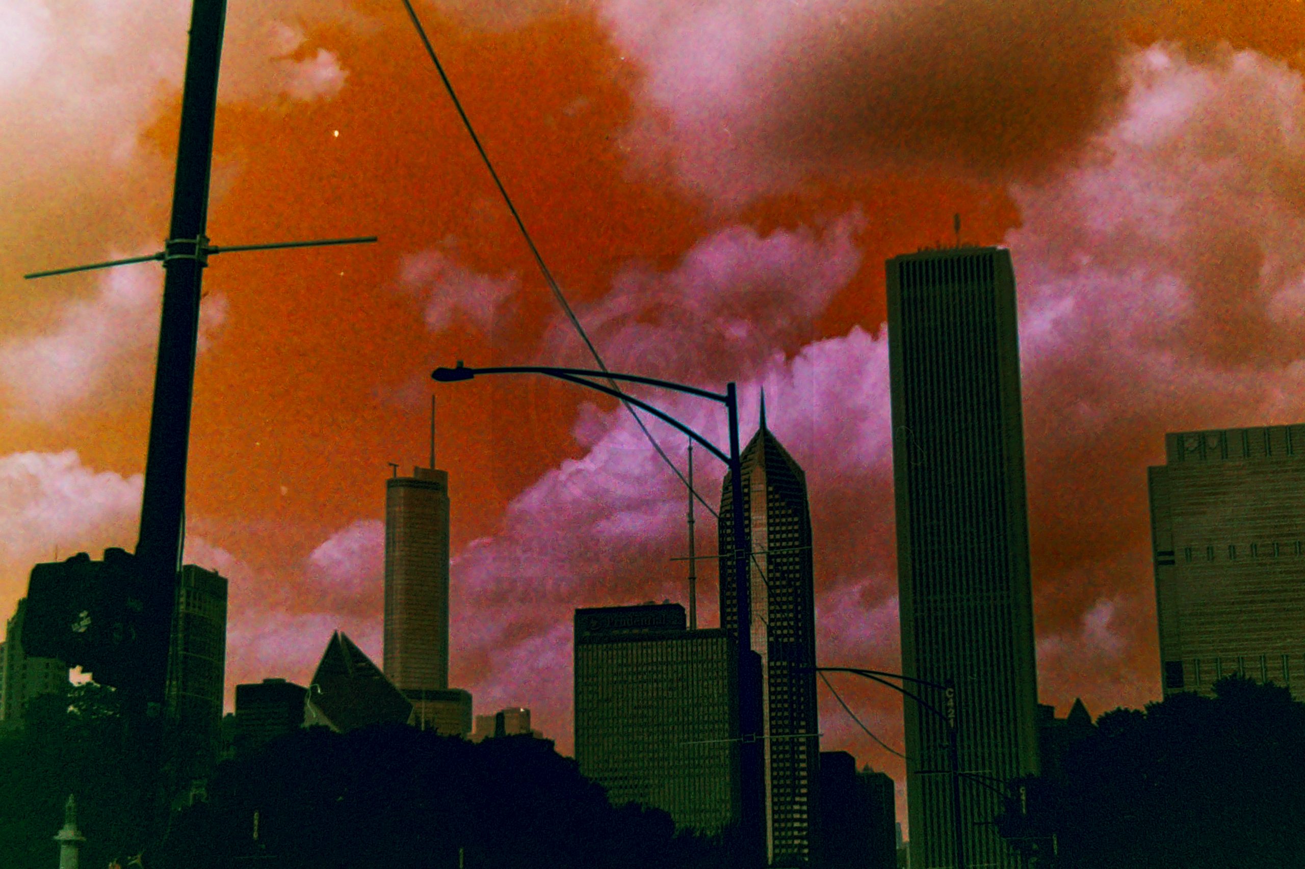 A low-angle shot of city skyscrapers silhouetted against a dramatic, surreal sky with vibrant orange and magenta clouds, captured with high-contrast, moody color shifts.